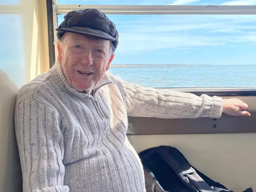 John Weeks sitting in a train window with blue sky and sea behind him, looking back smiling at the camera. Picture: Tracey Weeks