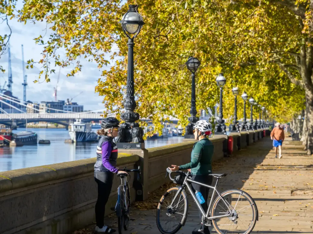 Two female cyclists wearing helmets and standing next to their bikes on a pedestrianised walk alongside the Thames in London with a bridge and skyscrapers in the background