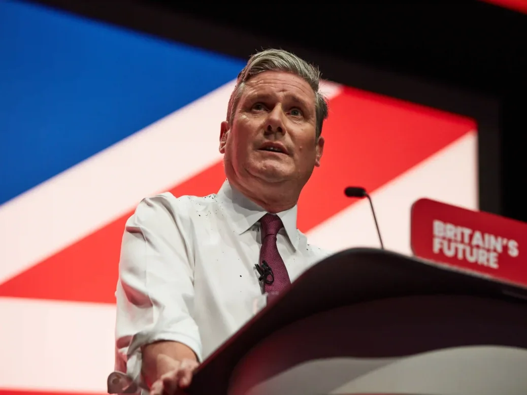 Sir Keir Starmer appears speaking to the 2023 Labour Party conference, sleeves rolled up and covered in glitter from a recent protest.