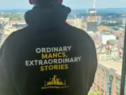 Person facing out of a tall window onto the Manchester skyline with the back of their hoodie reading 'Ordinary Mancs. Extraordinary stories' which is the new tagline for the website Manchester World